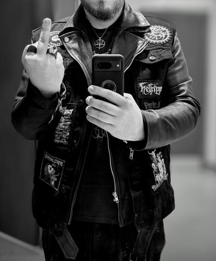 A black and white picture. A bearded gentleman wearing a black leather jacket with a sleeveless black denim jacket on top. The denim jacket has patches sewn on of various heavy metal bands. The man is also giving the camera the middle finger