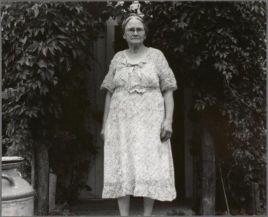 The image is a black and white photograph featuring an elderly woman standing in front of lush greenery, possibly bushes or vines. She has a stern expression on her face and wears a patterned knee-length dress with a bow detail around the neckline. Her hair appears to be short and she's wearing glasses. To her side stands a large ceramic jar or pot.

The background is filled with dense foliage that suggests an outdoor setting, perhaps in a garden or yard area of someone’s home. The lighting seems natural but slightly diffused, indicating either cloudy weather or shade from the surrounding vegetation.

There are no other people visible in the picture; the focus remains on this woman and her immediate surroundings. This image captures a moment frozen in time, presenting an intimate glimpse into the life of this individual during what appears to be late summer or early autumn based on the foliage's coloration and density.