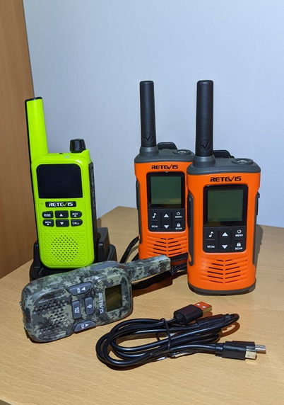 Several PMR446s. In the foreground a double USB cable, a camouflage RT33, on left a RA619 w- its charging cradle and on right two RT45Ps.