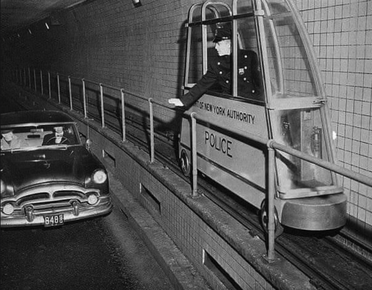 Black and white photo of one of the NYC under river tunnels. These tunnels connect the island of Manhattan to NJ and Brooklyn. A very narrow car that says "New York Authority Police" with a police officer signaling to a car on the road below the narrow tracks.