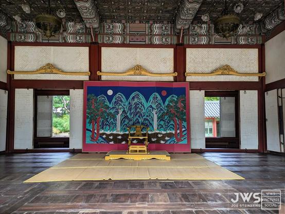 The image depicts a traditional Korean throne room, likely from the Joseon Dynasty (1392-1910). The room is decorated with intricate carvings and paintings on the walls and ceiling, and features a large, ornate throne at its center. The throne is adorned with gold accents and has a high back and armrests. In front of the throne is a low table or altar, which may be used for ceremonial purposes.

The room also contains several other pieces of furniture, including a large cabinet or screen on one wall and a small, intricately carved chair in the corner. The floor is made of dark wood and has a glossy finish, and there are several rugs or mats scattered throughout the room.

Overall, the image suggests that this is a formal, ceremonial space used for important events or rituals. The presence of the throne and altar, as well as the ornate decorations and furnishings, implies that this room was reserved for high-ranking officials or royalty.