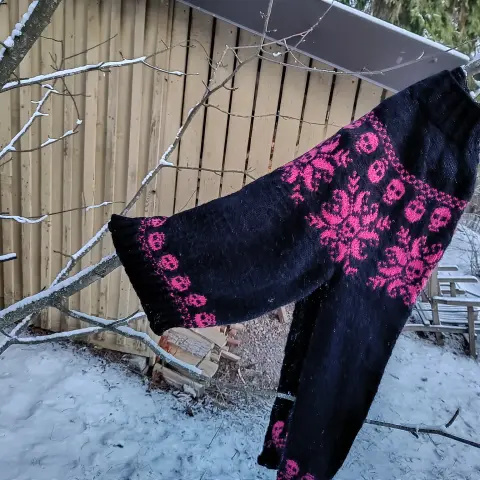 Kuvassa kaarrokeneule. Neuleen pohjaväri on musta. Neuleen kuviointi on pinkkiä. Neuleen  rintamusta kiertää lumihiutalekuvia, joka tarkemmin katsoen koostuu pääkalloista. Myös hihansuita ja helmaa kiertävät pääkallot.