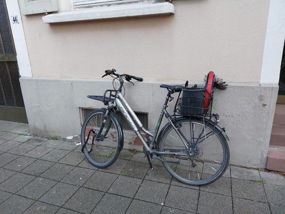 Ein "normales" Damenrad ist auf einem Gehweg abgestellt und lehnt an einer Hauswand. Auf dem Hinterradgepäckträger ist ein Korb mit Schornsteinfeger-Utensilien befestigt. Der Vorderradgepäckträger ist zum Zeitpunkt der Aufnahme ungenutzt.