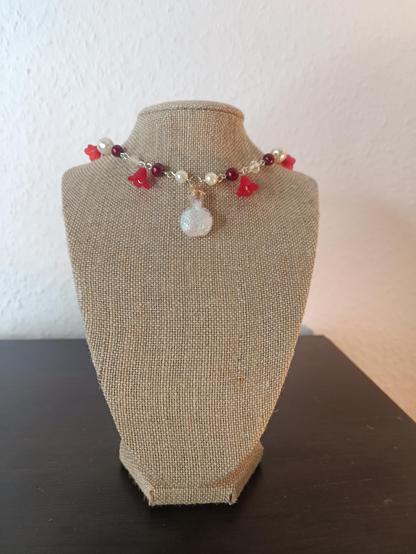 A necklace made of red beads, (fake) pearls and small red flowers. It is fitted tight to the neck. The pendant in the middle is a small bottle filled with iridescent strands.