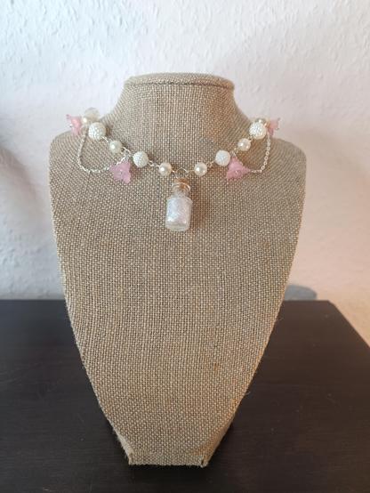 A necklace made of (fake) pearls and small pink flowers. It is fitted tight to the neck. The pendant in the middle is a small bottle filled with iridescent strands. Secrions of silver chain hang frm the sides.