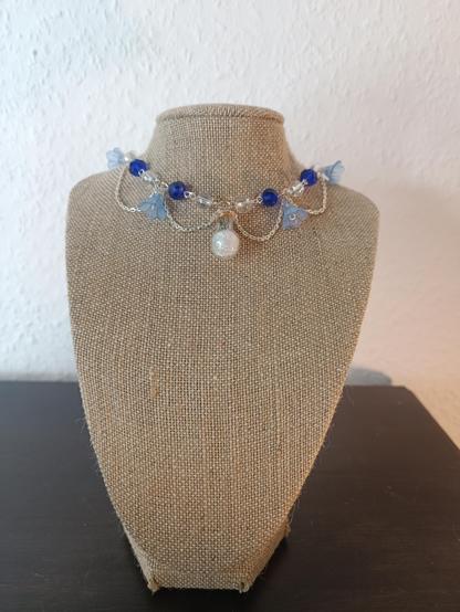 A necklace made of blue and clear beads and small blue flowers. It is fitted tight to the neck. The pendant in the middle is a small bottle filled with iridescent strands.