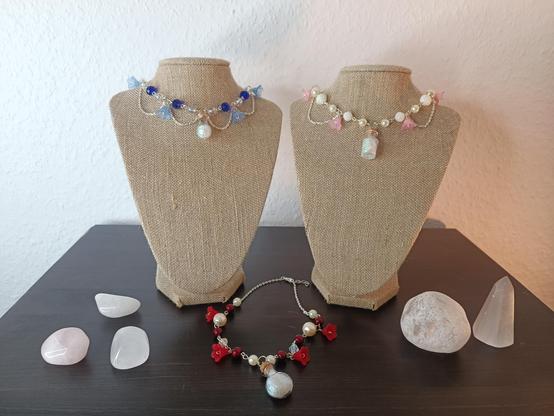 Two busts with necklaves stand on a table. On the left bust is a blue necklace. On the right one a mainly pearescent and pink one. Between the busts lies a red necklace. On the right and the left of the items lie several white stones.