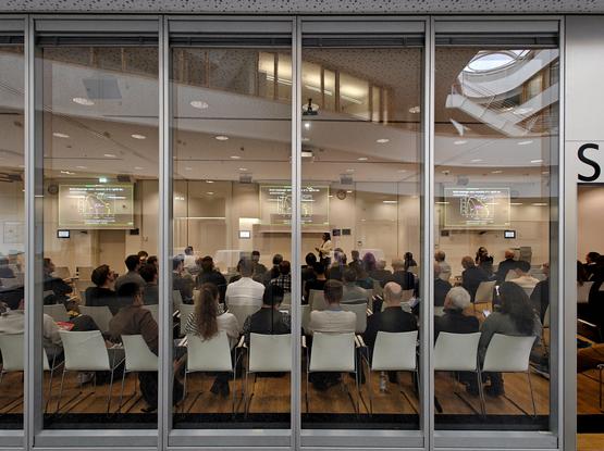This image shows a presentation in progress, viewed through glass panels. The room is filled with rows of white chairs occupied by attendees. At the front, there's a projection screen displaying a scientific graph. The space appears to be part of a larger modern building or facility, some details being reflected in the glass panels.