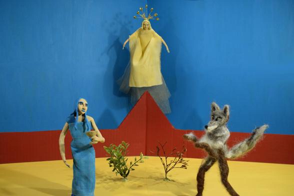 To the right, a claymation puppet wears blue and holds a gold goblet. 

In the center back of the image, a yellow puppet is suspended in the air above a red mountain. 

To the left, a needle felted wolf puppet looks at the blue puppet. 

The background is blue and the floor is yellow. There are two plants between the right and left puppet.