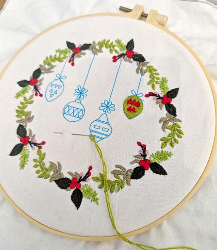 Embroidery hoop with a full embroidered christmas wrath in green tones. In the middle are printed baubles on the cloth which still need embroidery.