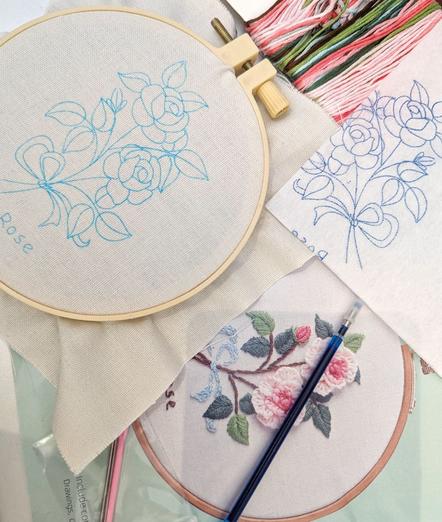 Small embroidery hoop with cloth. The cloth is printed with a rose pattern. Beneath is the embroidery silk, a ballpoint refill, fleece cloth with rose print and a photo of the finished embroidery.
