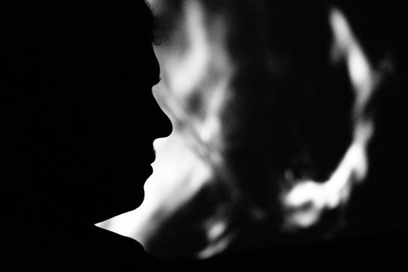 Das Bild zeigt eine Silhouette einer Person in einer Schwarz-Weiß-Komposition. Der Fokus liegt auf dem Profil des Gesichts, das durch den starken Kontrast zwischen Licht und Schatten klar umrissen ist. Die Person befindet sich im Vordergrund, vollständig in Dunkelheit gehüllt, wodurch die Konturen des Gesichts eine klare, minimalistische Wirkung erzeugen.

Im Hintergrund ist eine unscharfe, abstrakte Lichtstruktur sichtbar, die an Flammen oder Rauch erinnert. Die Formen im Hintergrund wirken diffus und weich, was der Szene eine mystische und geheimnisvolle Atmosphäre verleiht.

Die Kombination aus der scharfen Silhouette im Vordergrund und dem verschwommenen, leuchtenden Hintergrund erzeugt eine starke visuelle Spannung. Das Bild vermittelt Gefühle von Nachdenklichkeit oder Introspektion, wobei die künstlerische Darstellung Raum für Interpretation lässt. Die Einfachheit und der Kontrast zwischen Licht und Schatten verstärken die emotionale Tiefe der Szene.