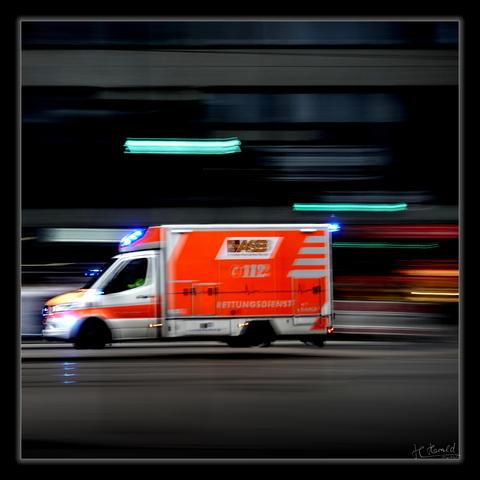 Square image with a black frame of a bright red ASB ambulance with blue lights in action. The vehicle drives from right to left through the image and is slightly blurred due to the panning effect used. Green, red and orange horizontal light stripes can be seen in the dark, very blurred background.