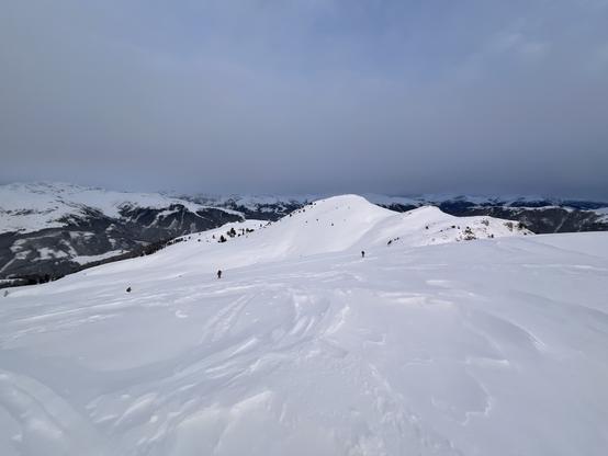 Three splitboarders in the distance, hiking up to a plateau, one of numerous false summits on the way to Kirchspitze. Distant mountains are lightly covered by a hazy layer of cloud which would soon burn away as the sun came up. The ground is slightly wind scoured snow, but quite deep. It is somewhere around 8am.