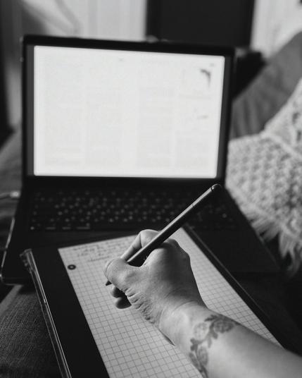 A black and white photo of a hand writing with a pen on a tablet with grid paper. A floral tattoo is visible on the wrist. In the background, an open laptop displays blurred text on the screen. The person is sitting on a couch or a soft chair, with a knitted pillow nearby.
