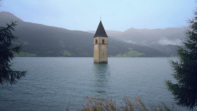 A tall, slender church steeple rises from a lake, with mountains and a cloudy sky in the background.
