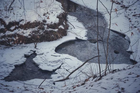 A creek in winter. The creek bends through the frame, the water rushing through the thick blanket of snow on either side of it.