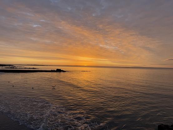 A sunrise over the sea. The sky is various hues of orange and blue. The surface of the water is peppered with waves and sea foam.
