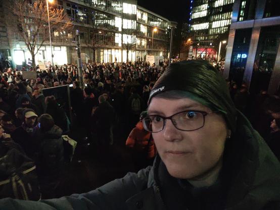 Frau mit Mütze und Brille im Vordergrund, dahinter eine Menschenmenge.