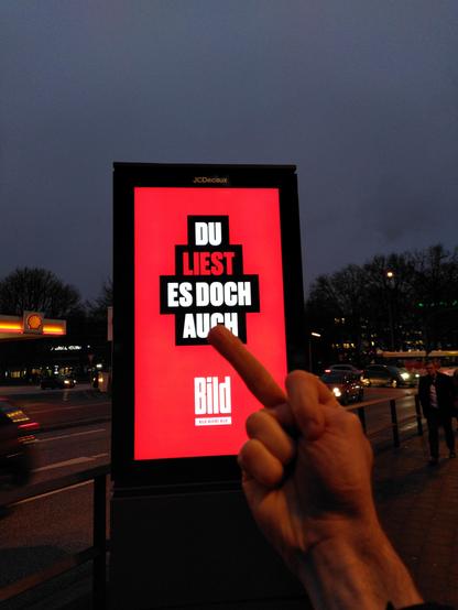 LED-Anzeige mit Werbung für BILD: "Du liest es doch auch! BILD" Im Vordergrund des Fotos streckt der Fotograf dieser Anzeige einen ausgestreckten Mittelfinger entgegen.