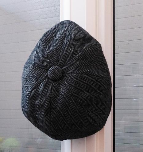 Casquette de type gavroche en tweet, motif en chevron gris, accrochée à la poignée d' une fenêtre moderne. derrière, un volet roulant blanc baissé