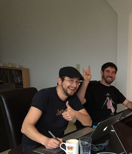 Zwei kaukasische Männer in schwarzen T-Shirts sitzen vor Laptops und lachen in die Kamera