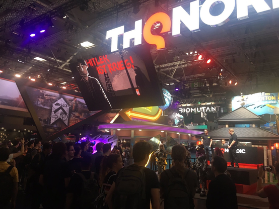 a crowded hall at Gamescom and a huge screen showing a scene from the game where somene paints the slogan "Hitler ist Krieg" - "Hitler means war" at a awall
