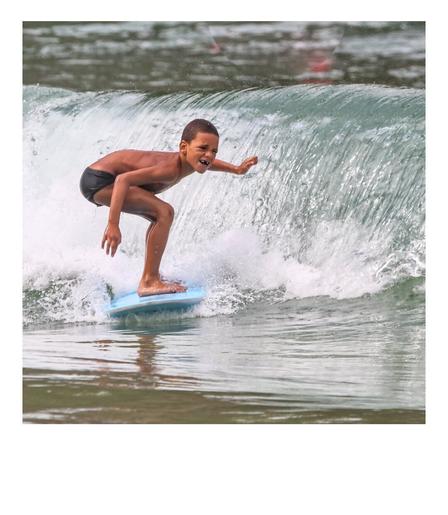 A very young tooth-gapped surfer boy riding a wave standing up on his blue board. His smile shows a funny open window :)