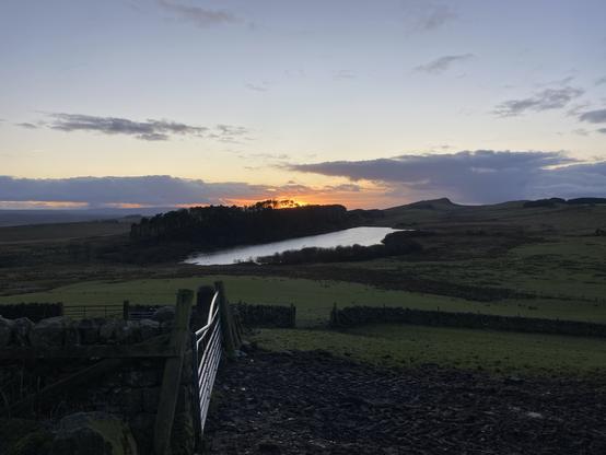 A landscape at sunset, taken from a farmyard with a farm gate visible in the foreground, and the sun going down behind a wide ridge with a lake visible in front of it