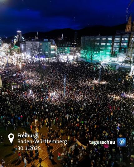 Eine riesige Demonstration in der Innenstadt von Freiburg, Baden-Württemberg, am 30. Januar 2025. Es wurde am Abend aufgenommen, der Himmel ist dunkelblau, und die Stadtbeleuchtung erhellt die Straßen. Tausende von Menschen haben sich auf einem großen Platz versammelt, der von modernen Gebäuden mit beleuchteten Fenstern umgeben ist.

Die Menge ist dicht gedrängt, viele Teilnehmende halten Schilder und Banner hoch, die jedoch aus der Distanz nicht lesbar sind. Einige Bereiche der Demonstration sind besonders hell erleuchtet, möglicherweise durch Scheinwerfer oder Kameras. Die Straßen und Gebäude in der Umgebung sind ebenfalls gut beleuchtet, einige Fassaden sind grün angestrahlt.

In der unteren linken Ecke des Bildes befinden sich weiße Texteinblendungen mit Informationen zur Stadt (Freiburg, Baden-Württemberg) und zum Datum (30.1.2025). In der unteren rechten Ecke befindet sich das Logo der „Tagesschau“, was darauf hinweist, dass das Bild von einem Nachrichtenmedium stammt.

Insgesamt vermittelt das Bild eine kraftvolle, dynamische Atmosphäre einer Großdemonstration mit beeindruckender Beteiligung.