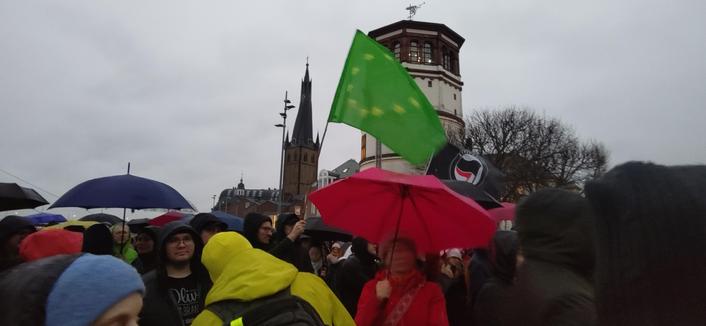 Demonstrierende mit Regenschirmen vor dem Turm des Schifffahrtsmuseums und dem von St. Lambertus in Düsseldorf. Zu sehen ist auch eine grüne Fahne mit kreisförmig angeordneten gelben Sternen, wie bei der Europafahne.