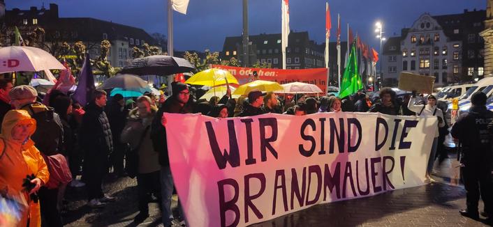 Demonstrierende, teilweise mit Regenschirmen, sammeln sich hinter einem großen Transparent mit der Aufschrift "WIR SIND DIE BRANDMAUER".