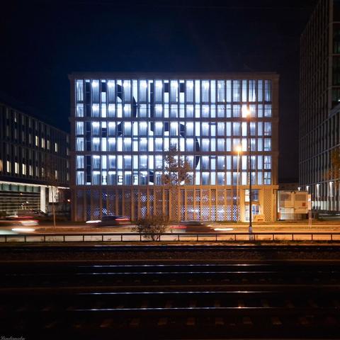 Cubusförmiges, blau beleuchtetes Gebäude bei Nacht mit Schienen im Vordergrund.