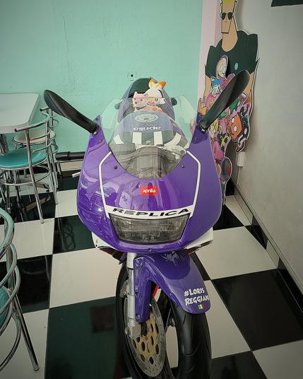 A purple Aprilia Replica motorcycle is parked on display in a room with a black and white checkered floor. The motorcycle has a stuffed animal, a white cat with orange spots, sitting on the gas tank. There is a cartoon cutout of Johnny Bravo and Dexter's Laboratory characters in the background. To the left of the motorcycle are teal diner-style tables and chairs.