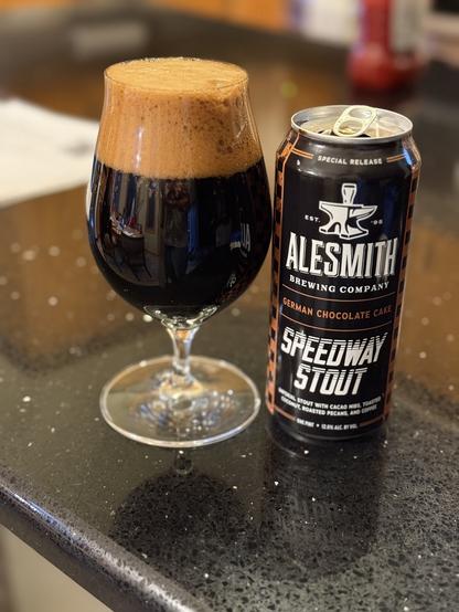 A snifter glass full to the very brim with dark beer and a huge dark tan head. The can is black with white lettering and brown accents. The Alesmith anvil logo is front and center