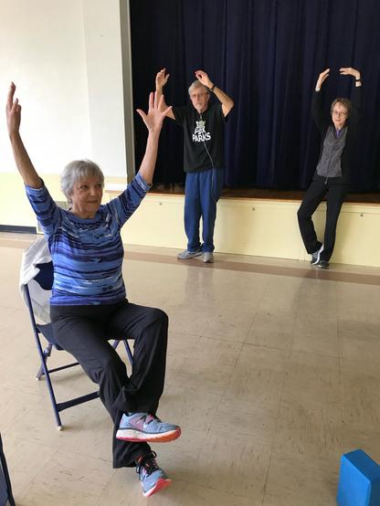 Three older adults doing a yoga tree pose. A argue woman with silver hair is doing a seated version in the foreground. In the background are two people doing standing versions against the edge of a stage. On the right is another white woman with dark hair, to the left is a white man using an oxygen tank.