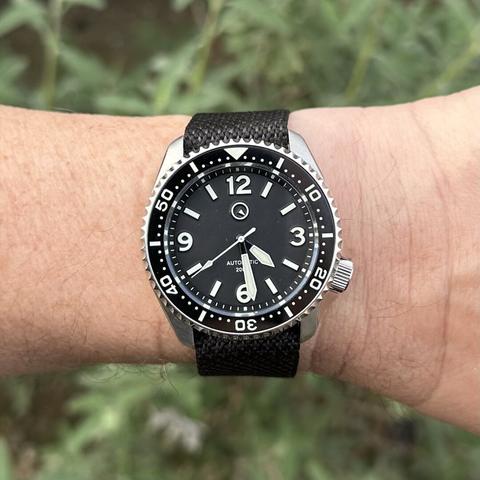 Watch with a brushed stainless steel case, 4:00 crown, black dial with numeric indices at 12, 3 , 9, and 6, baton indices elsewhere, white lume-filled sword hands, and black ceramic dive bezel with white numerals and markers, on a black fabric strap.