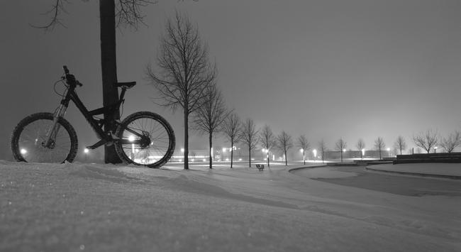 Black and white photo of my black Cannondale Super-V bike with silver Lyric suspension fork (12 or 16 cm travel) and Rohloff hub on the completely snow-covered Avantis terrain between Kerkrade, Netherlands, and Aachen, Germany at night. The background is formed by trees on a road that is a has circular curve and a concentric watercourse and by street lights.