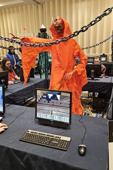 A mannequin demon in an orange robe stands in the middle of multiple tables. On the table in front of the demon there's a monitor, keyboard, and mouse. DOOM is running on the computer.