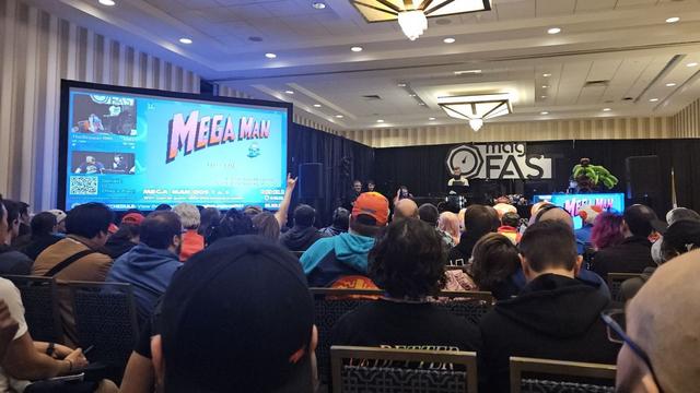 A large screen showing the main menu for Mega Man DOS 1; a stage with several people and a piano; and a crowd of people watching.