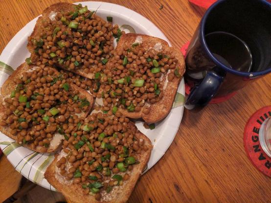 Four slices of bread on a paper plate with tahina, lentils, and green onion. Green tea to the side.