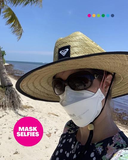 A person taking a selfie on a sunny beach, wearing a wide-brimmed straw hat with a Roxy logo, large dark sunglasses, and a white 3M Aura N95 respirator. The beach has white sand, palm trees, and a calm ocean in the background. A pink circle in the bottom left corner contains the text "MASK SELFIES." At the top, six colorful dots are arranged in a row.