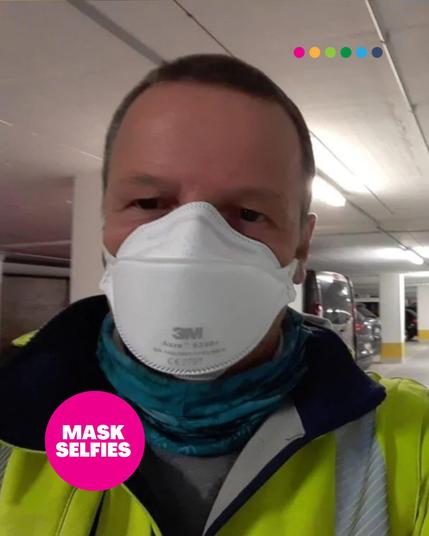 A person taking a selfie in an indoor parking garage, wearing a high-visibility yellow jacket, a blue neck gaiter, and a white 3M Aura N95 respirator. The garage is well-lit with cars visible in the background. A pink circle in the bottom left corner contains the text "MASK SELFIES." At the top, a row of six colorful dots is visible.