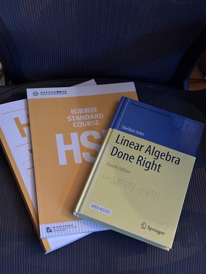 A fanned-out stack of text books. The top book is a yellow hardback volume with a blue stripe at the top, entitled “Linear Algebra Done Right”. Underneath it is an orange softcover course book with a white stripe at the top and a grey-and-white striped stripe at the bottom. It is titled “Standard Course HSK 1”. Above the English title, a line of Chinese hanzi say the same. Underneath this second book is another softcover with the same color scheme but flipped. The title is obscured; it is a workbook for the characters required by the HSK 1 proficiency exam.