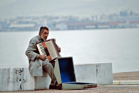 A disheveled man in grey busks with a piano accordion, its blue-fabric-lined case yawning invitingly, on low rectangular formations near an urban waterfront area.