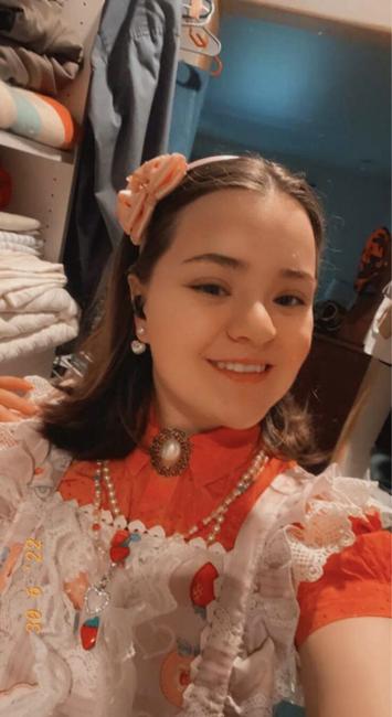 Selfie of Dolly in her Lolita jump skirt only shown from the chest up. In this she wears a lilac colored dress skirt with white laced trim, strawberry and tea set graphics as well as a row of deceptive strawberry buttons
On the bodice. That combined with a red, short sleeved blouse which is pinned at the collar with a pearl cameo pendant, two pearl necklaces. 

Along to match the necklace’s are pearl heart earrings, a lilac headband decorated with pink bows
