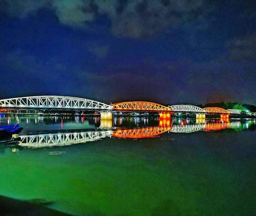 Photo shows an iron bridge made in wrought iron style from the early 1900s. It is light by multicolor lamps in each section. This colours here are white, orange, white red and green. The background is deep blue sky, and the river is deep green.