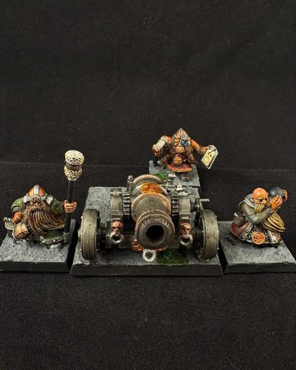A Warhammer dwarf cannon crew. A steel cannon is surrounded by three crew. One holds a cannonball ready to load. A second consults a clipboard while adjusting the aim. The third stands by with a sponge stick, a tankard of ale in his other hand.