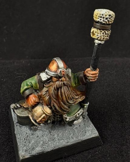 A Warhammer dwarf cannon crew miniature. A dwarf with a flowing brown beard wears a green tunic and a copper cap rimmed in steel. He stands next to a lidded iron bucket holding a long pole with a sponge attached to one end. He stands ready to clean the barrel of his weapon once it fires. In his other hand is a gilded mug of ale. A bit of refreshment while he waits to do his job.