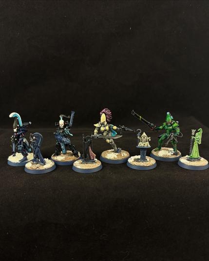 Four Warhammer Eldar aspects warrior miniatures stand next to their associated shrine miniatures. A Dire Avenger in dark blue armor with a feathered helmet holds a rifle standing by a grey stone figure decorated with gems. A black armored Dark Reaper with a skull helm aims a long gun next to an onyx figure of a shrouded reaper. A Howling Banshee in grey and bone armor with flowing purple hair wields a sword beside a small white stone figure on a pedestal. Finally, a Striking Scorpion in green and brown armor wields a chainsword beside a jade figure outfitted with the same equipment.